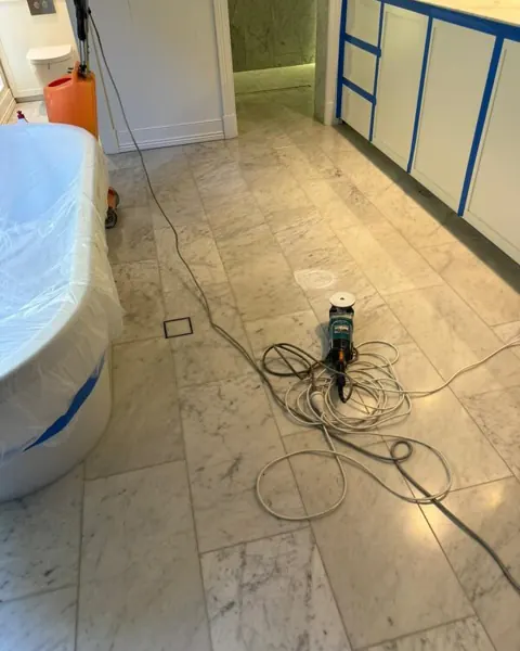 White marble bathroom floor with a grinder and cables on it, the bath and cabinets masked with blue tape