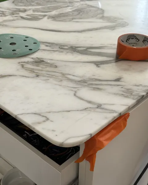 White marble benchtop with dramatic grey veining, a green polishing pad on top and orange tape along the front edge
