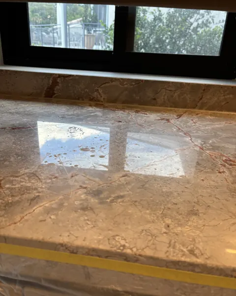 Tan marble benchtop after polishing, reflecting the window frame, with masking tape still on the wall